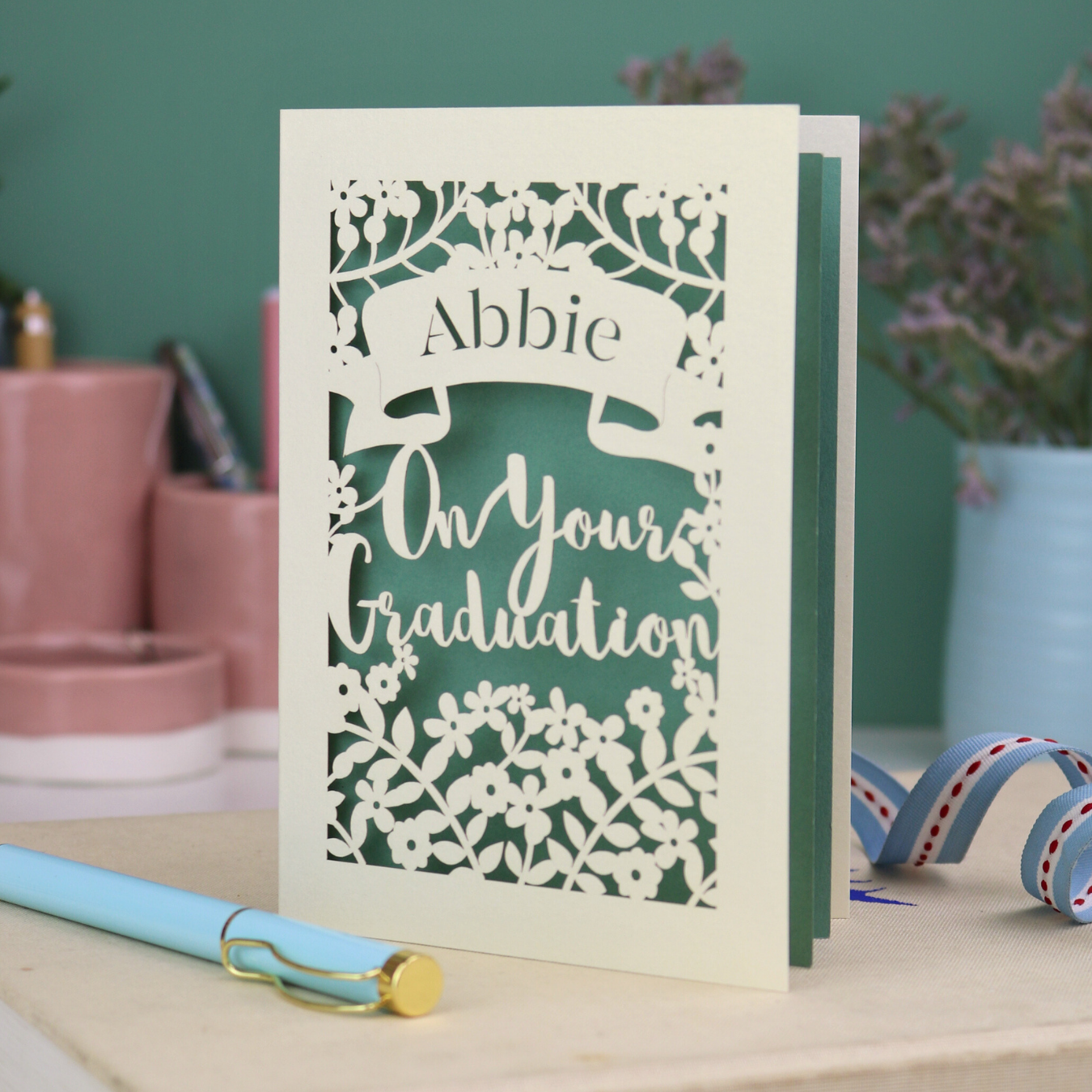 Laser cut Graduation card with personalized name 'Abbie' and floral cut out design, on a table with decorative elements.