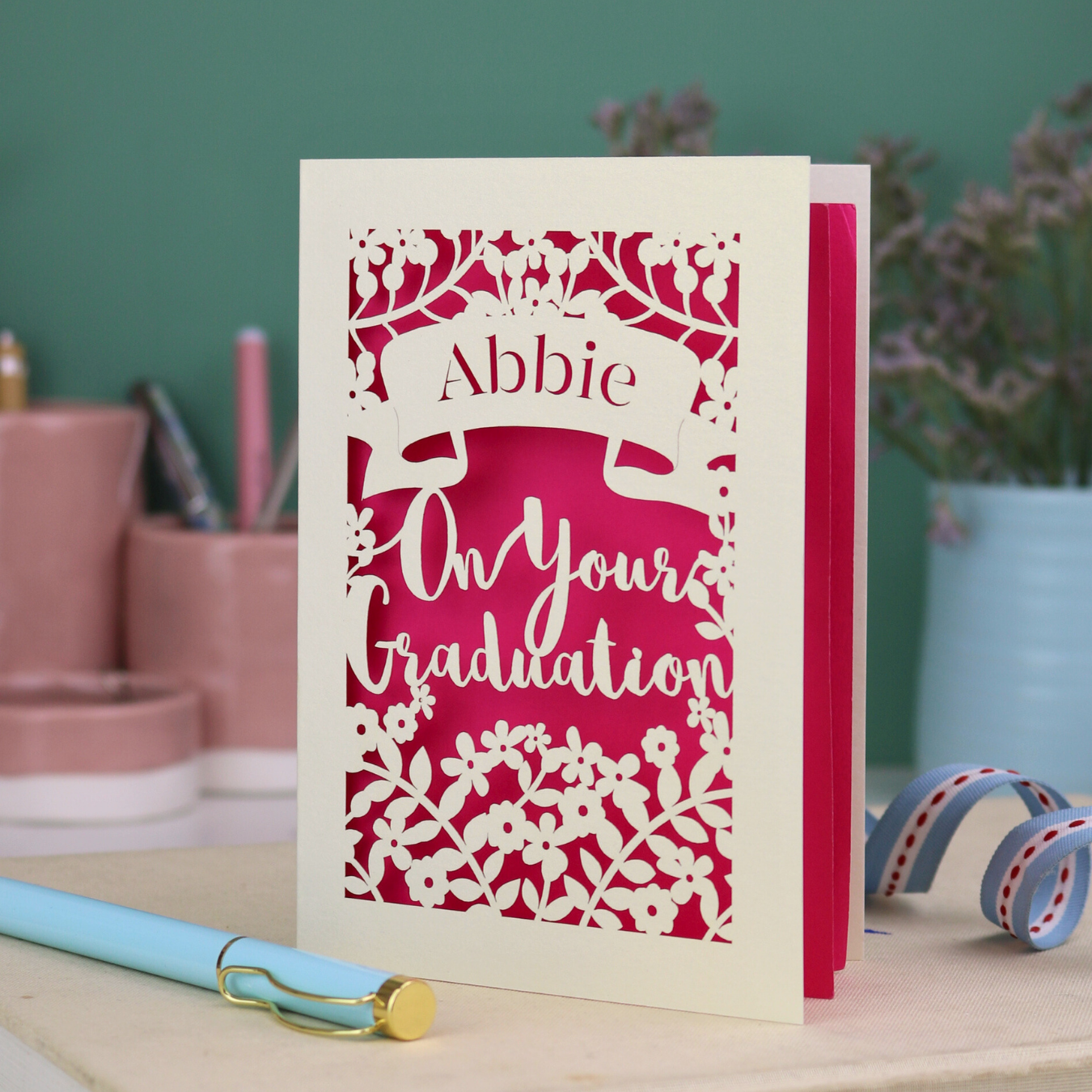 Laser cut Graduation card with personalized name 'Abbie' and floral cut out design, on a table with decorative elements.