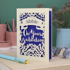 Laser cut Graduation card with personalized name 'Abbie' and floral cut out design, on a table with decorative elements.