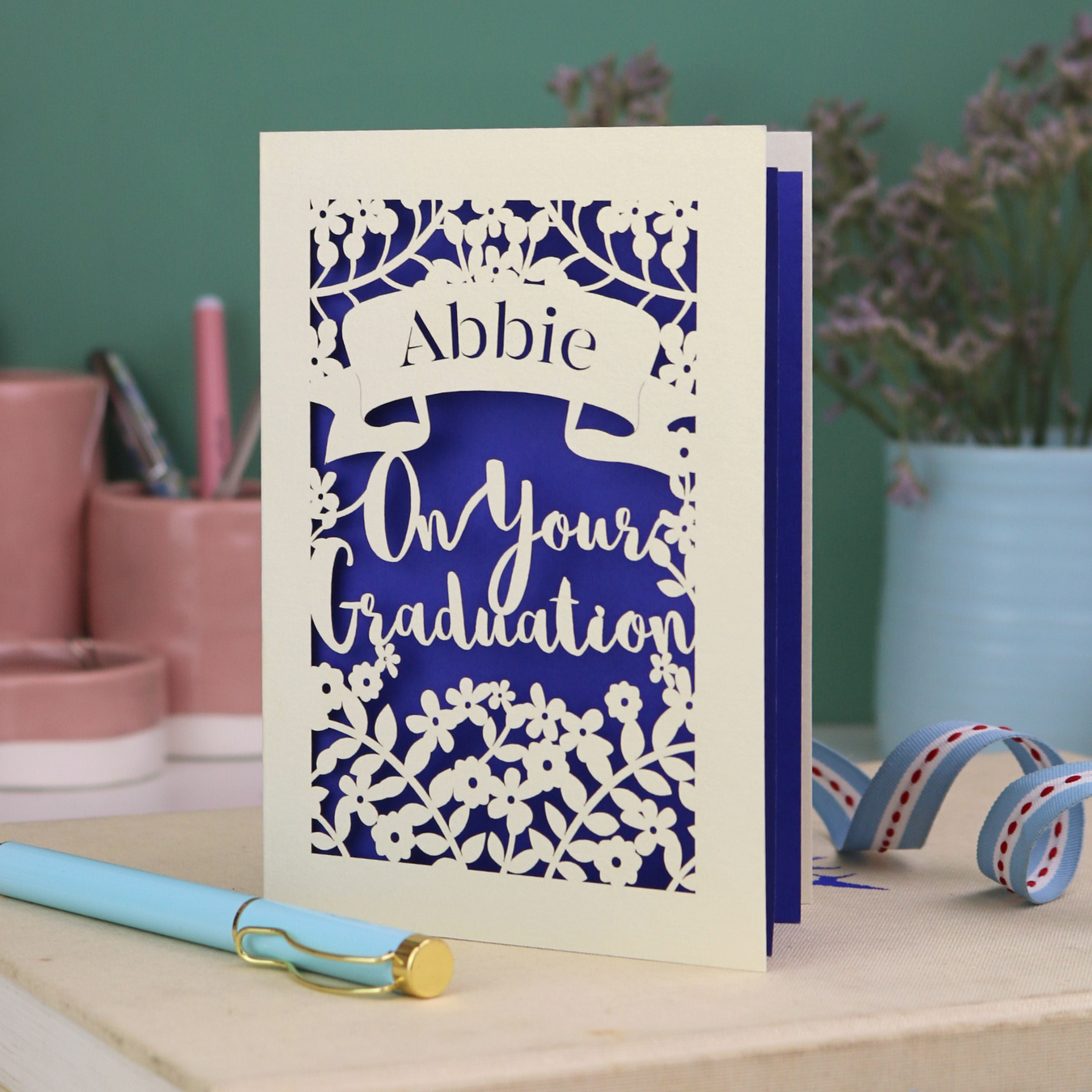 Laser cut Graduation card with personalized name 'Abbie' and floral cut out design, on a table with decorative elements.