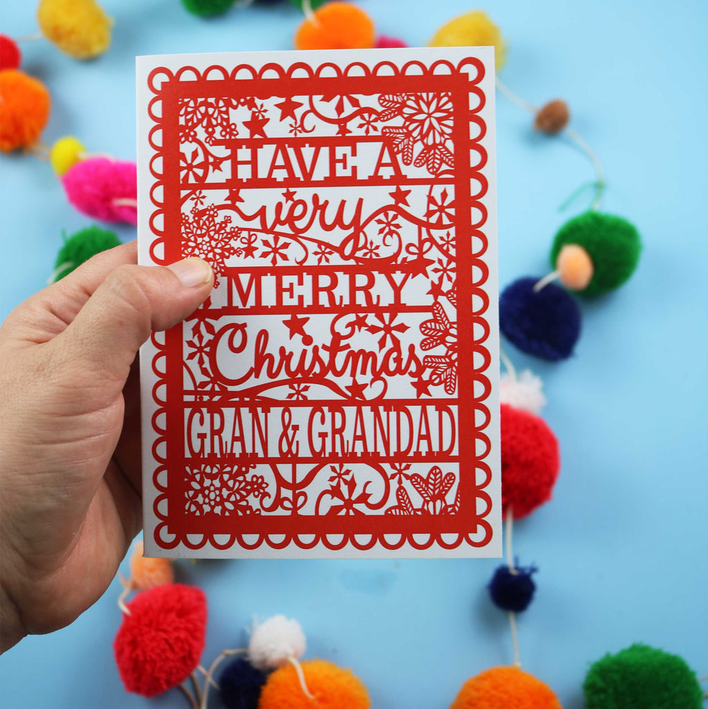 Hand holding a red and white Christmas card with personalized text against a colourful pom-pom background.