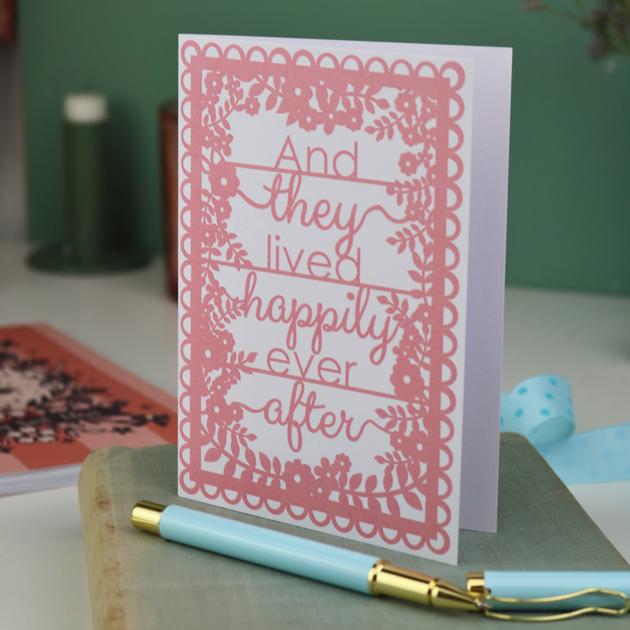 Pink printed card with 'And they lived happily ever after' text on a white surface with a pen and decorative items.