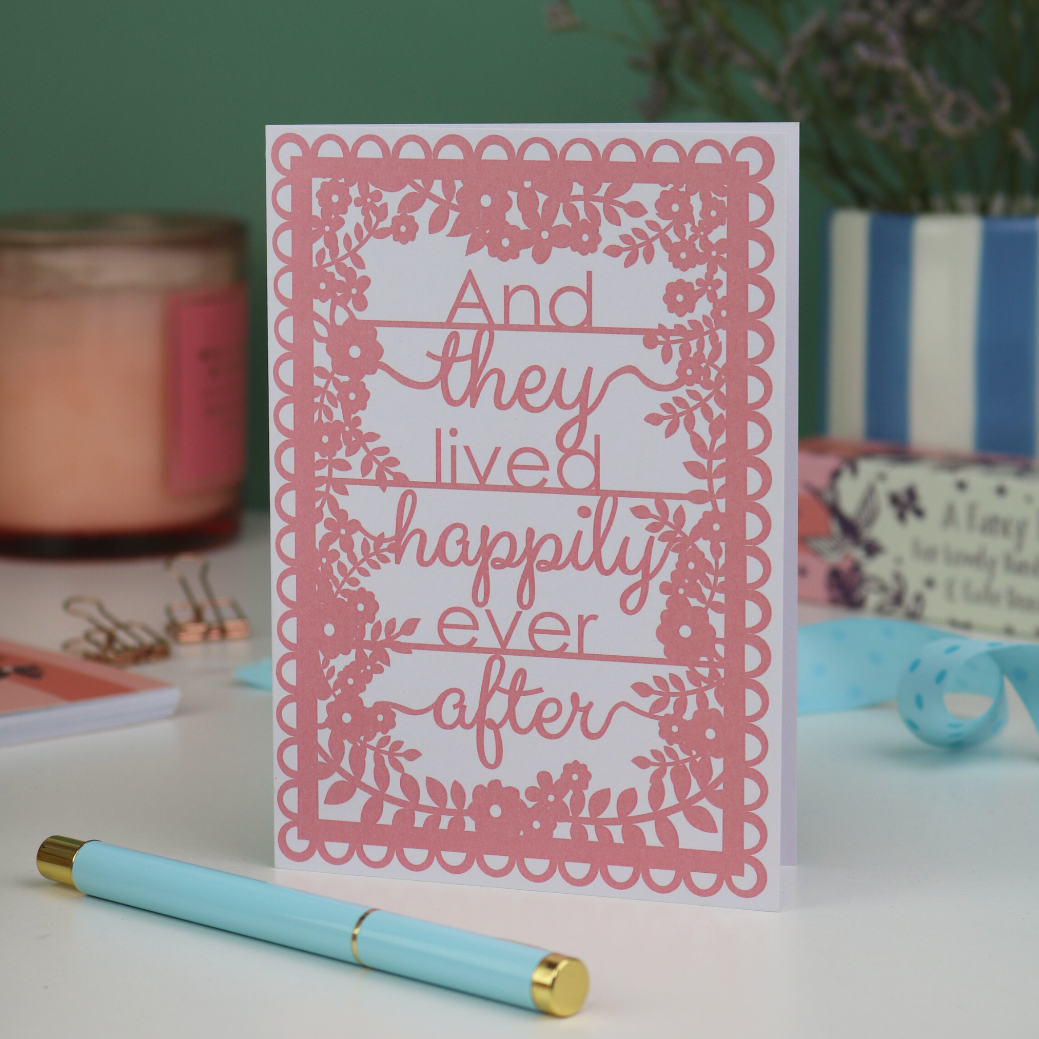 Pink printed card with 'And they lived happily ever after' text on a white surface with a pen and decorative items.