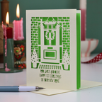 A green and cream laser-cut Christmas card featuring a front door with a wreath, brick walls, and the text 'Rob, Carly and Olive Happy 1st Christmas in your new home'. The card is displayed on a white surface with a pen in the foreground and blurred candles in the background.