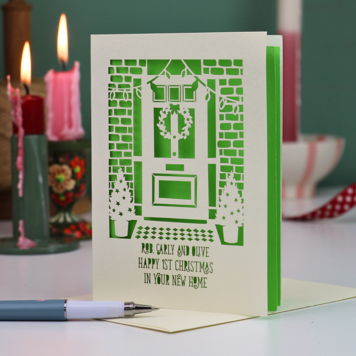 A green and cream laser-cut Christmas card featuring a front door with a wreath, brick walls, and the text 'Rob, Carly and Olive Happy 1st Christmas in your new home'. The card is displayed on a white surface with a pen in the foreground and blurred candles in the background.