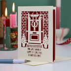 A Christmas card for a new home, with a laser-cut design of a front door decorated with a wreath, set on a tabletop with blurred pink candles and holiday décor in the background.