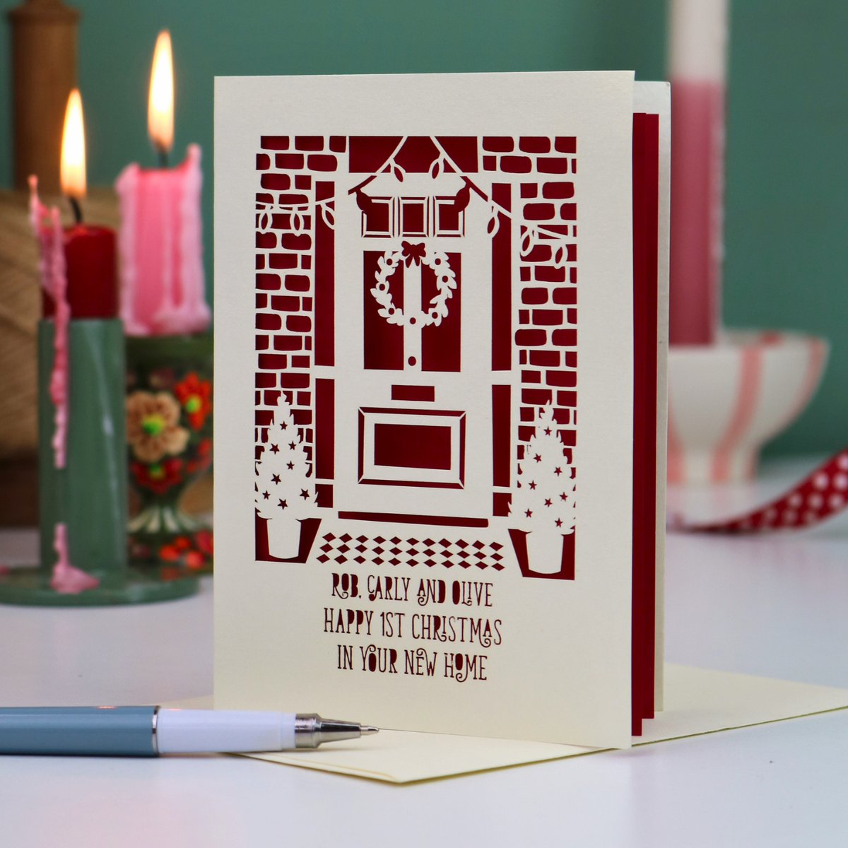 A Christmas card for a new home, with a laser-cut design of a front door decorated with a wreath, set on a tabletop with blurred pink candles and holiday décor in the background.