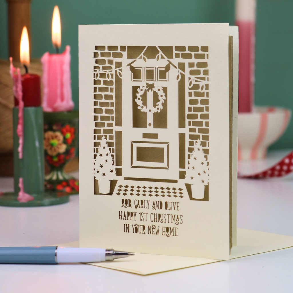 A cream-colored personalised laser-cut greeting card featuring a festive Christmas front door design with wreath, potted trees, and the message 'Rob, Carly and Olive, Happy 1st Christmas in your new home'. The card is displayed on a light surface with a pen in the foreground and blurred pink candles in the background.