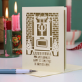 A cream-colored personalised laser-cut greeting card featuring a festive Christmas front door design with wreath, potted trees, and the message 'Rob, Carly and Olive, Happy 1st Christmas in your new home'. The card is displayed on a light surface with a pen in the foreground and blurred pink candles in the background.