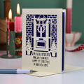 A handmade Happy first Christmas in your new home card with a violet backing and laser-cut illustration of a front door, wreath, brick wall, and potted plants, displayed on a table with candles in the background.