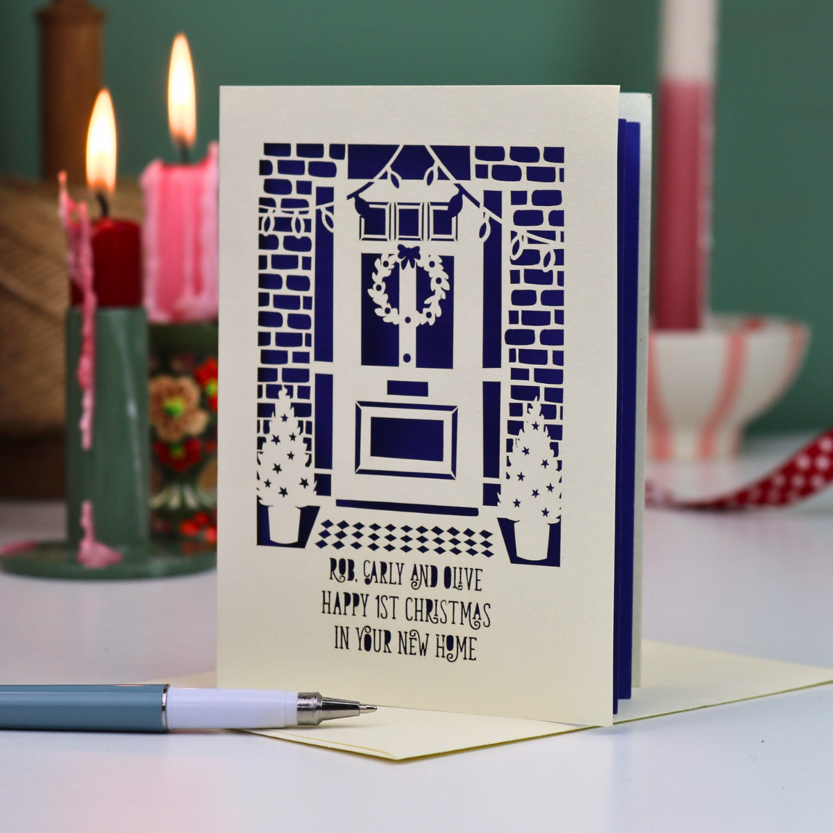 A handmade Happy first Christmas in your new home card with a violet backing and laser-cut illustration of a front door, wreath, brick wall, and potted plants, displayed on a table with candles in the background.