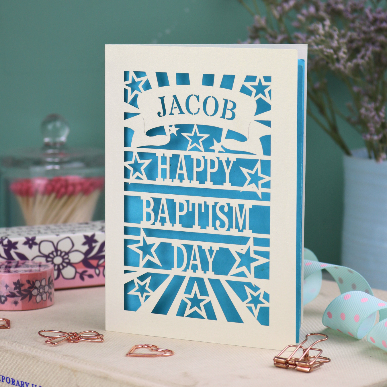 Personalised Laser Cut Greeting card with 'Happy Baptism Day' text on a decorative surface with flowers and stationery items.