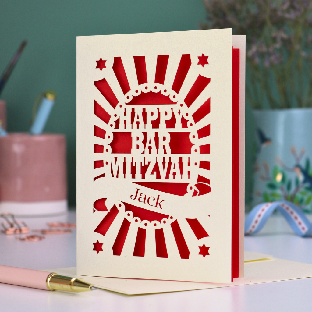Red and cream laser cut Bar Mitzvah card with 'Happy Bar Mitzvah Jack' text on a desk with stationery items.