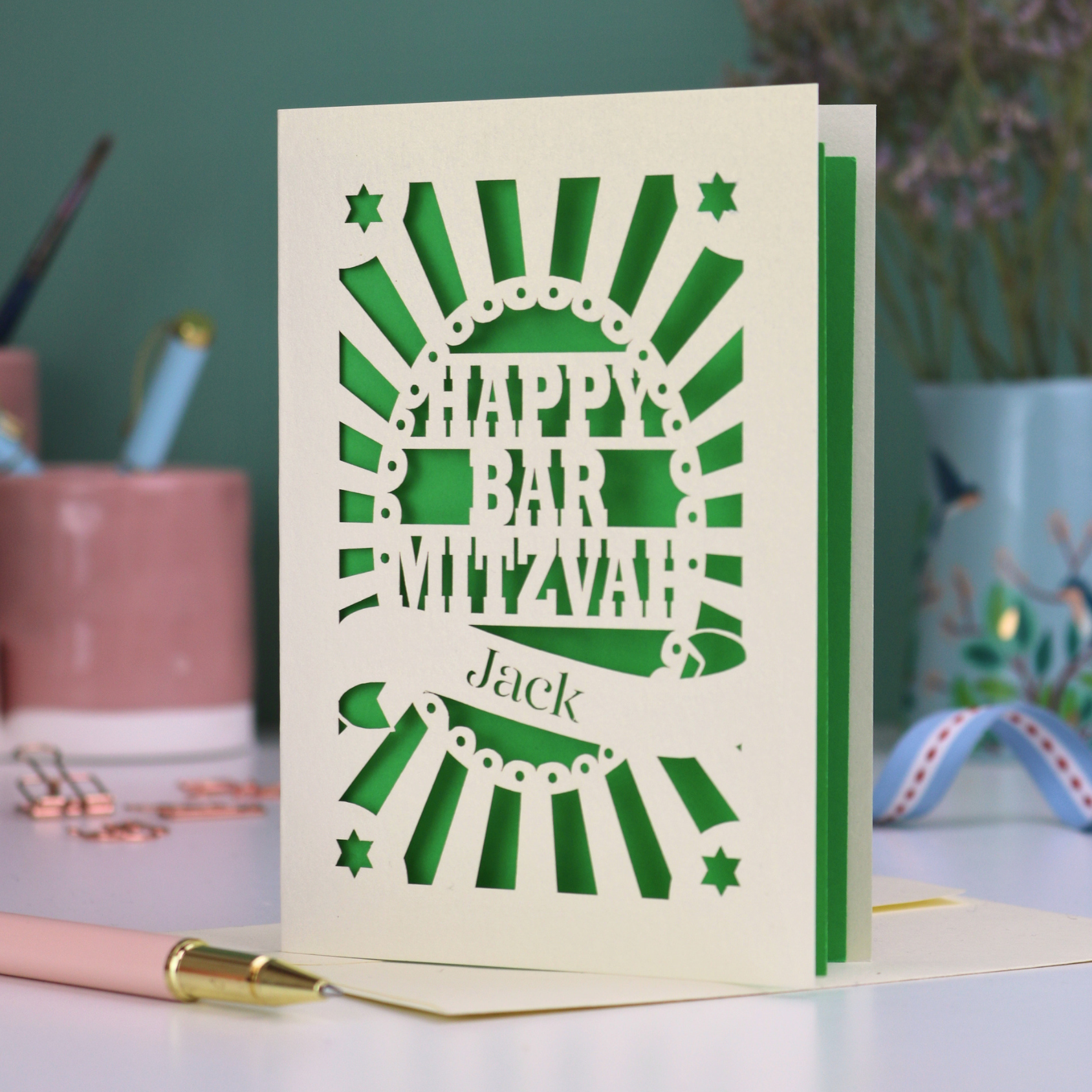 Green and cream laser cut Bar Mitzvah card with 'Happy Bar Mitzvah Jack' text on a desk with stationery items.