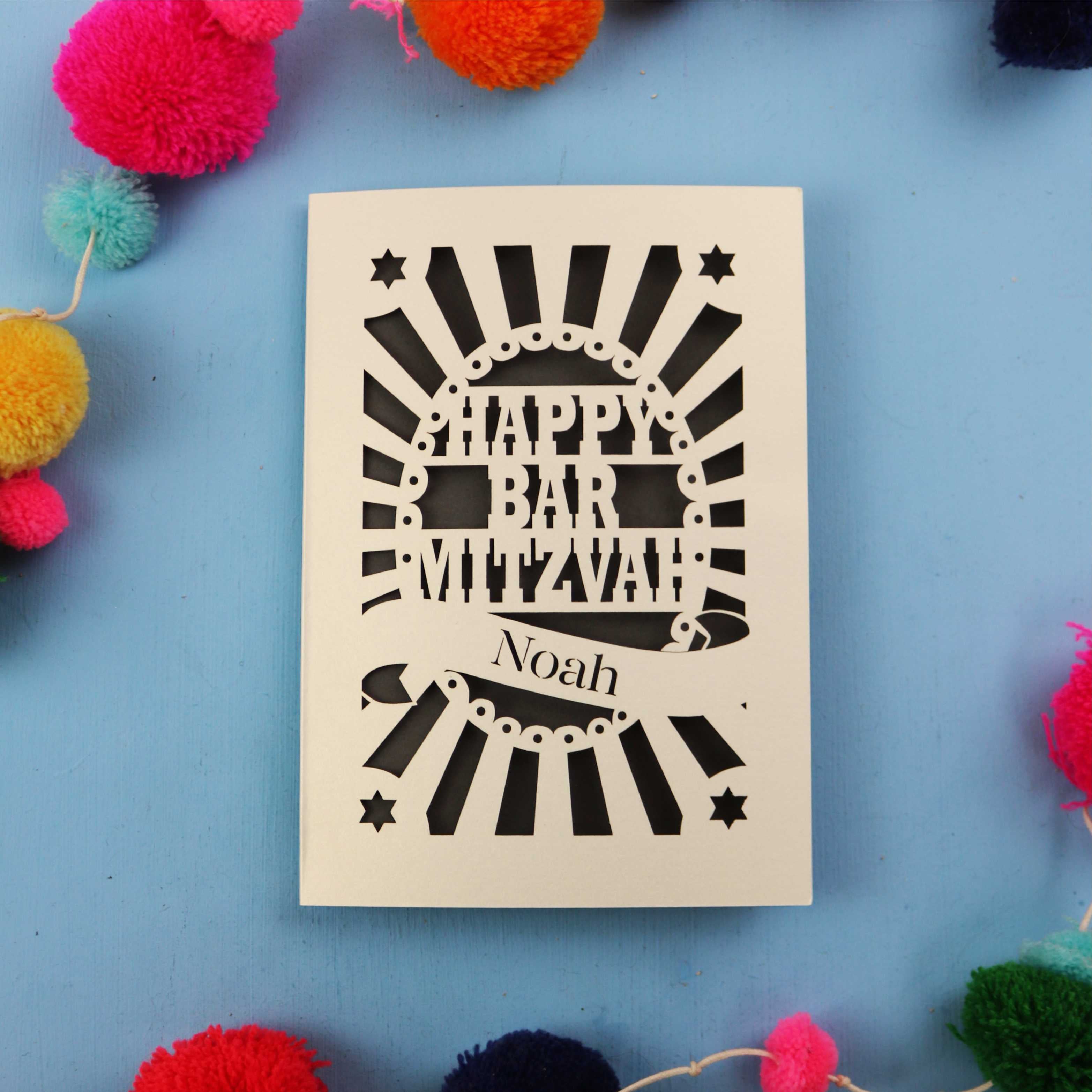 Laser Cut card with 'Happy Bar Mitzvah Noah' text on a blue background with colourful pom-poms.