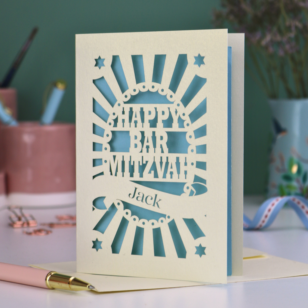 Blue and cream laser cut Bar Mitzvah card with 'Happy Bar Mitzvah Jack' text on a desk with stationery items.