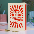 Orange and cream laser cut Bar Mitzvah card with 'Happy Bar Mitzvah Jack' text on a desk with stationery items.