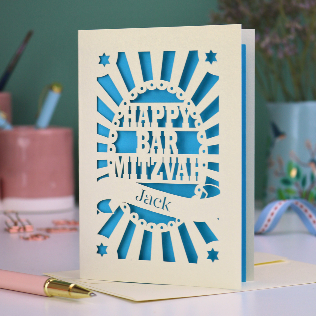 Blue and cream laser cut Bar Mitzvah card with 'Happy Bar Mitzvah Jack' text on a desk with stationery items.
