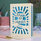 Blue and cream laser cut Bar Mitzvah card with 'Happy Bar Mitzvah Jack' text on a desk with stationery items.