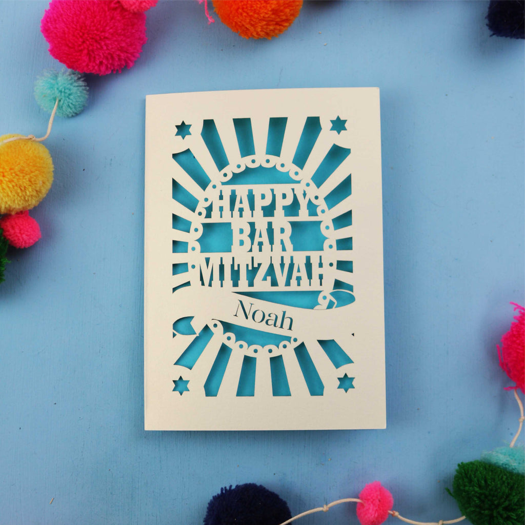 Laser Cut card with 'Happy Bar Mitzvah Noah' text on a blue background with colourful pom-poms.