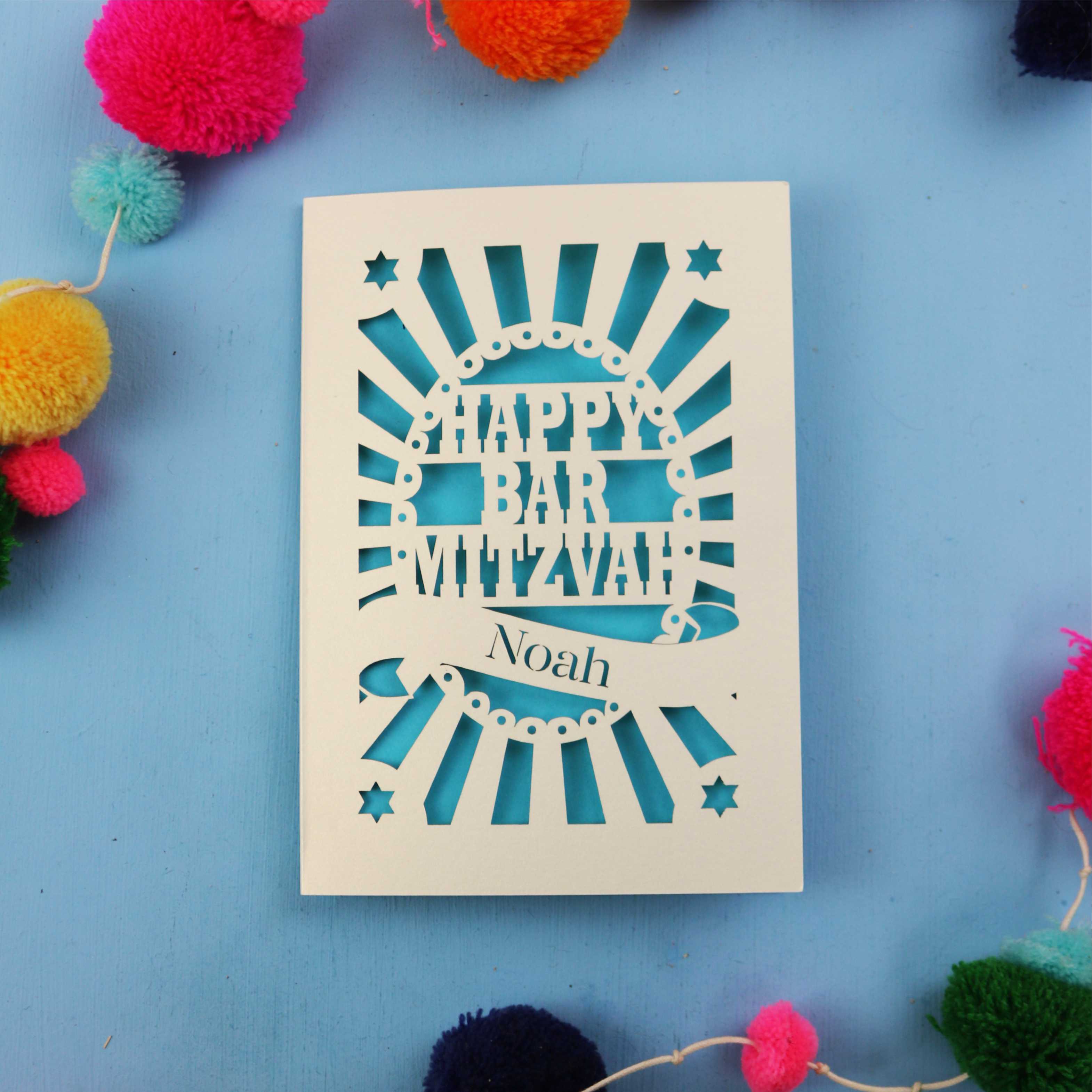 Laser Cut card with 'Happy Bar Mitzvah Noah' text on a blue background with colourful pom-poms.
