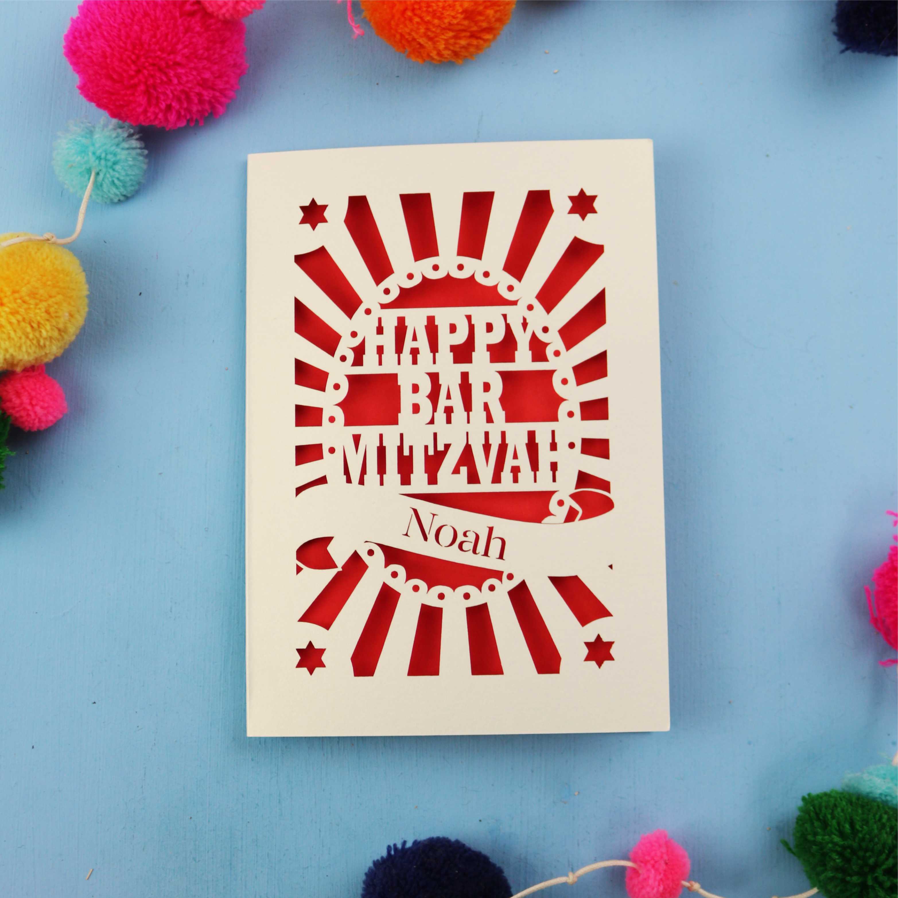 Laser Cut card with 'Happy Bar Mitzvah Noah' text on a blue background with colourful pom-poms.