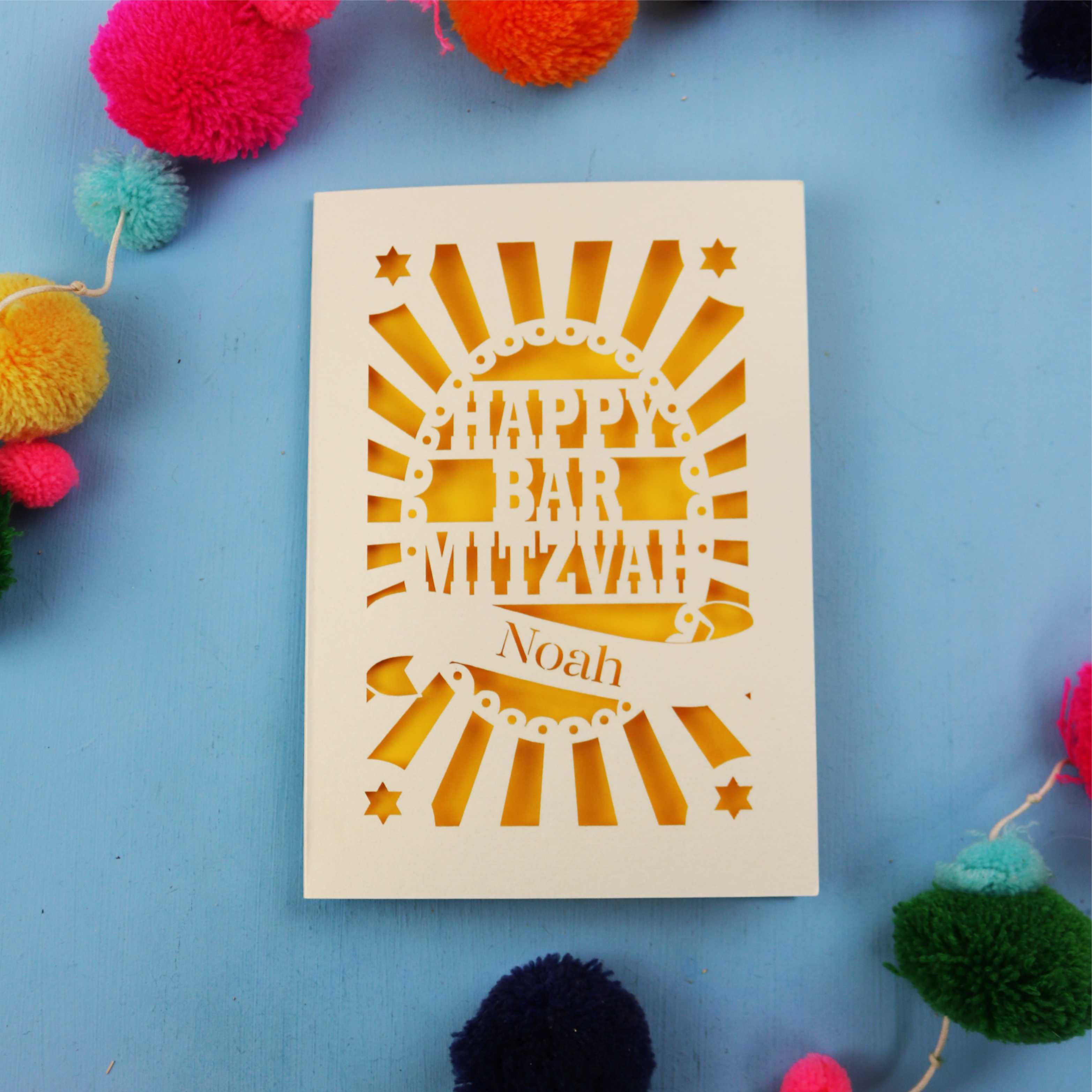 Laser Cut card with 'Happy Bar Mitzvah Noah' text on a blue background with colourful pom-poms.