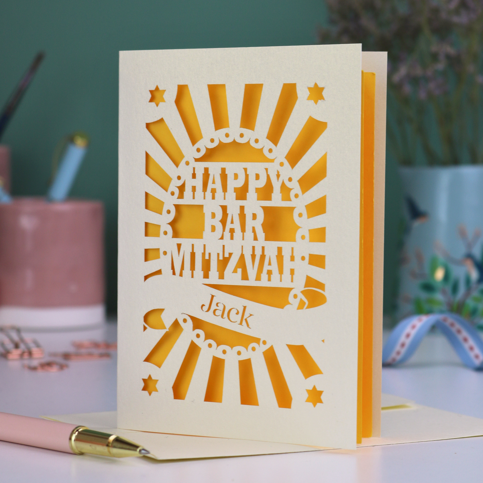 Yellow and cream laser cut Bar Mitzvah card with 'Happy Bar Mitzvah Jack' text on a desk with stationery items.