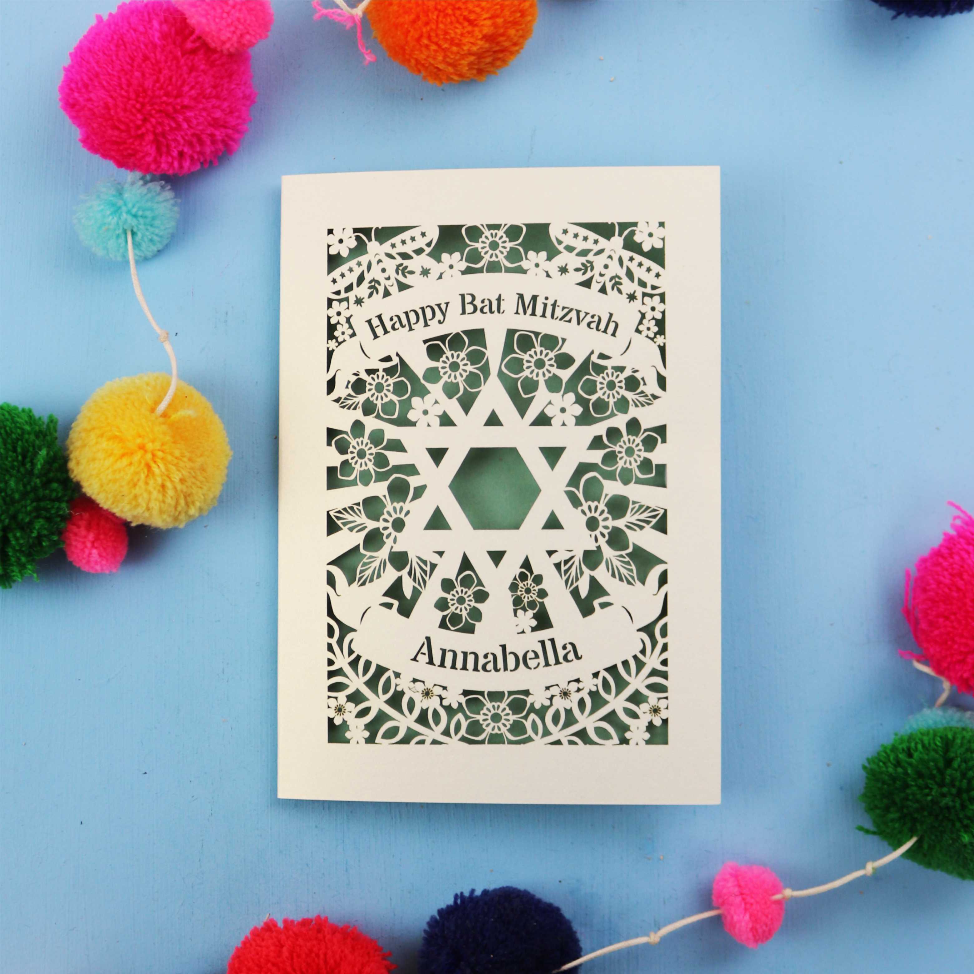 Card with 'Happy Bat Mitzvah' and 'Annabella' text on a blue background with colourful pom-poms.