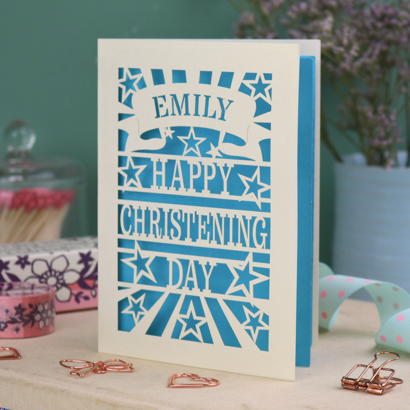 Personalized laser cut christening card with blue background, and text, "Emily, Happy Christening Day", with star design, placed on a surface with decorative items.