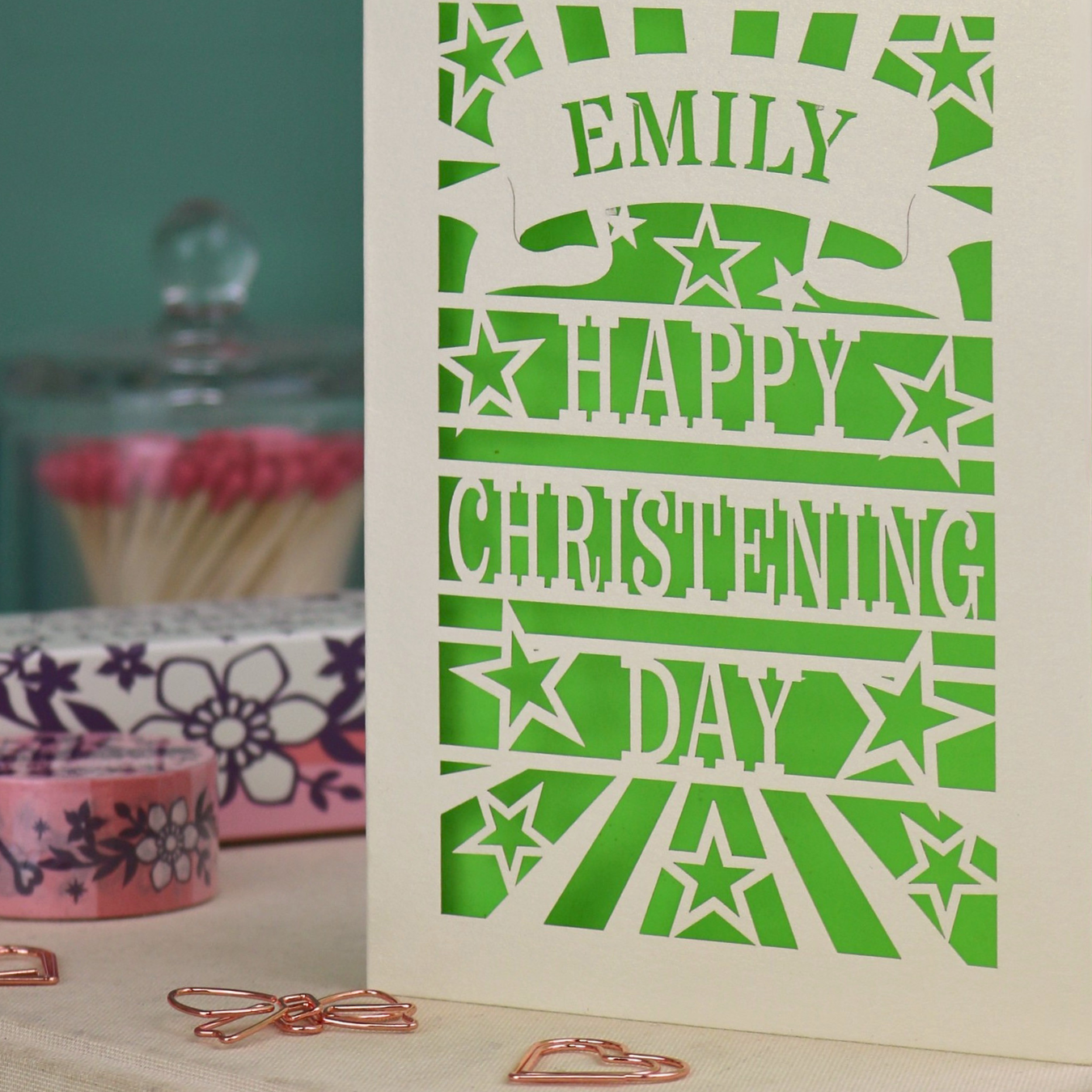 Personalized laser cut christening card with green background, and text, "Emily, Happy Christening Day", with star design, placed on a surface with decorative items.