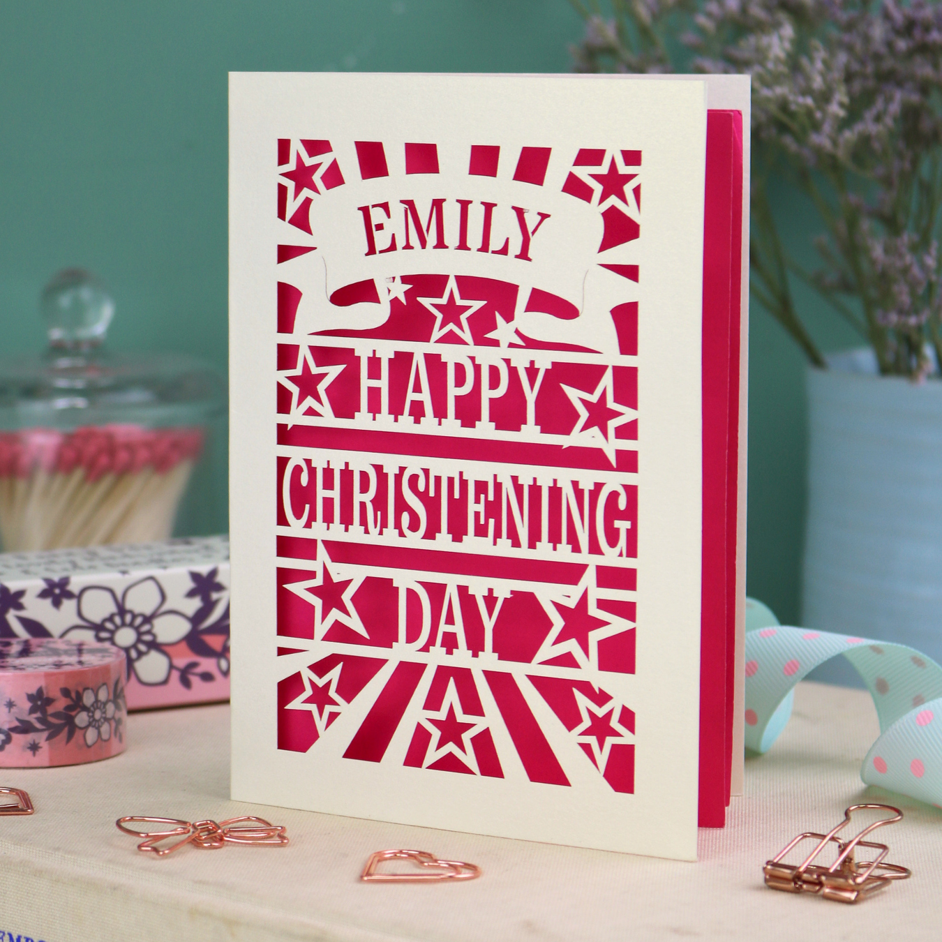 Personalised laser cut christening card with shocking pink background, and text, "Emily, Happy Christening Day", with star design, placed on a surface with decorative items.