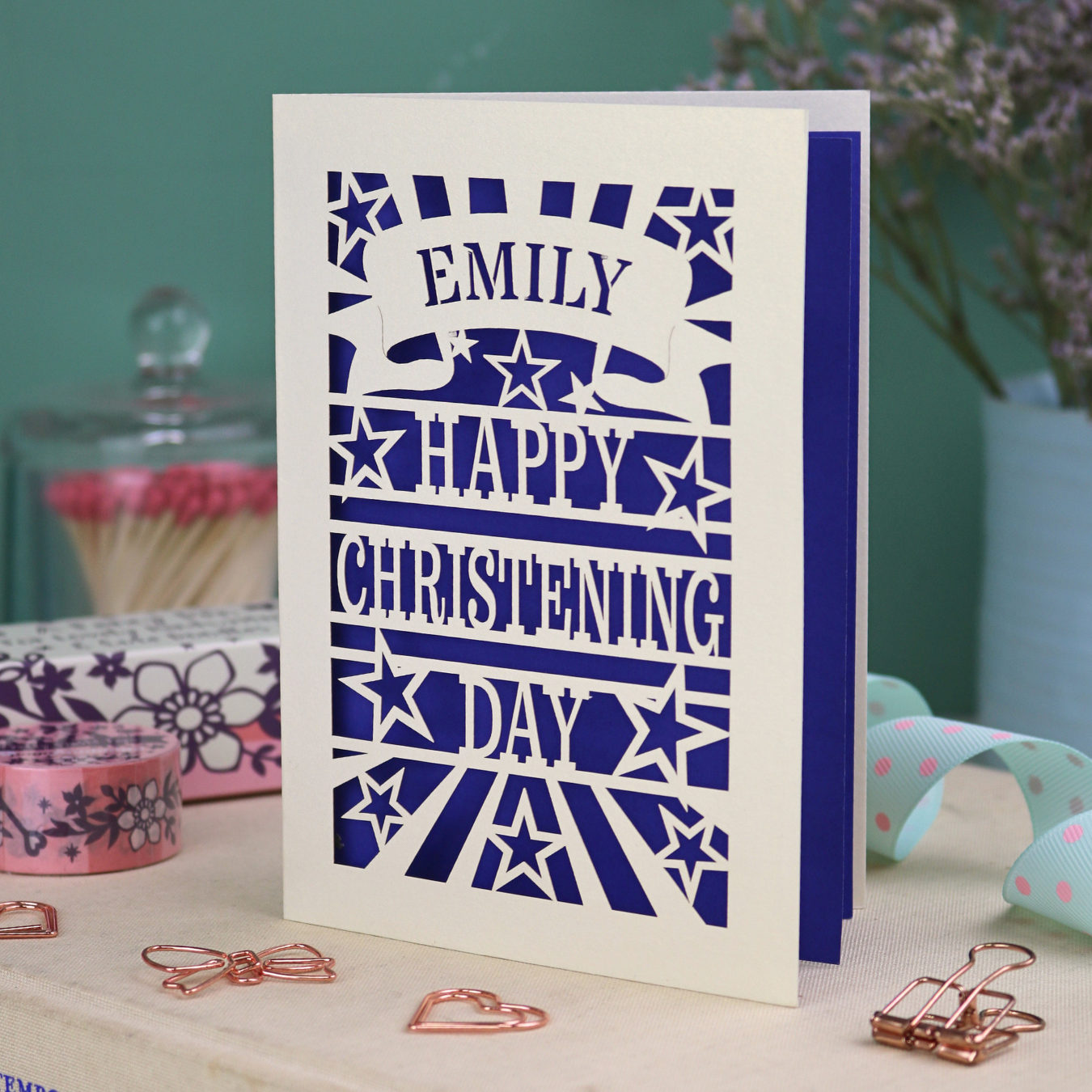 Personalized laser cut christening card with violet background, and text, "Emily, Happy Christening Day", with star design, placed on a surface with decorative items.