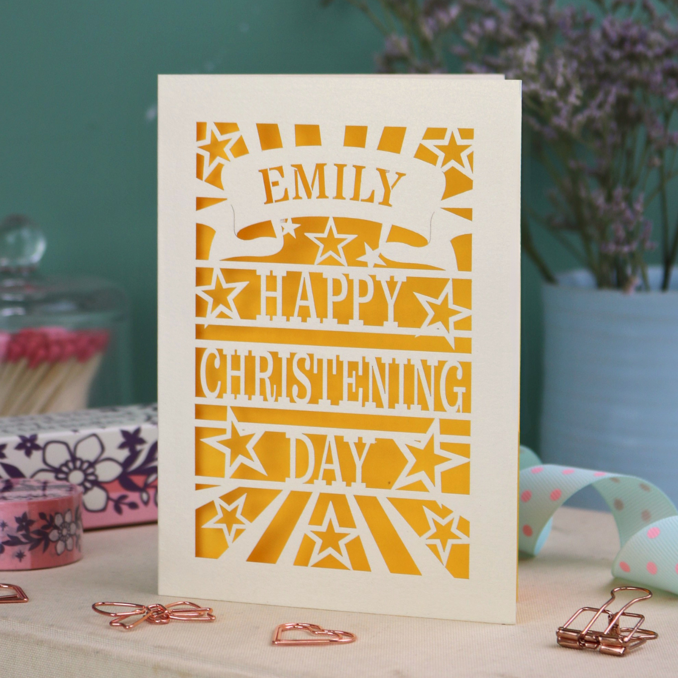Personalized laser cut christening card with yellow background, and text, "Emily, Happy Christening Day", with star design, placed on a surface with decorative items.