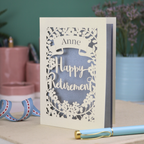 Laser-cut 'Happy Retirement' card with personalized name on a decorative surface.