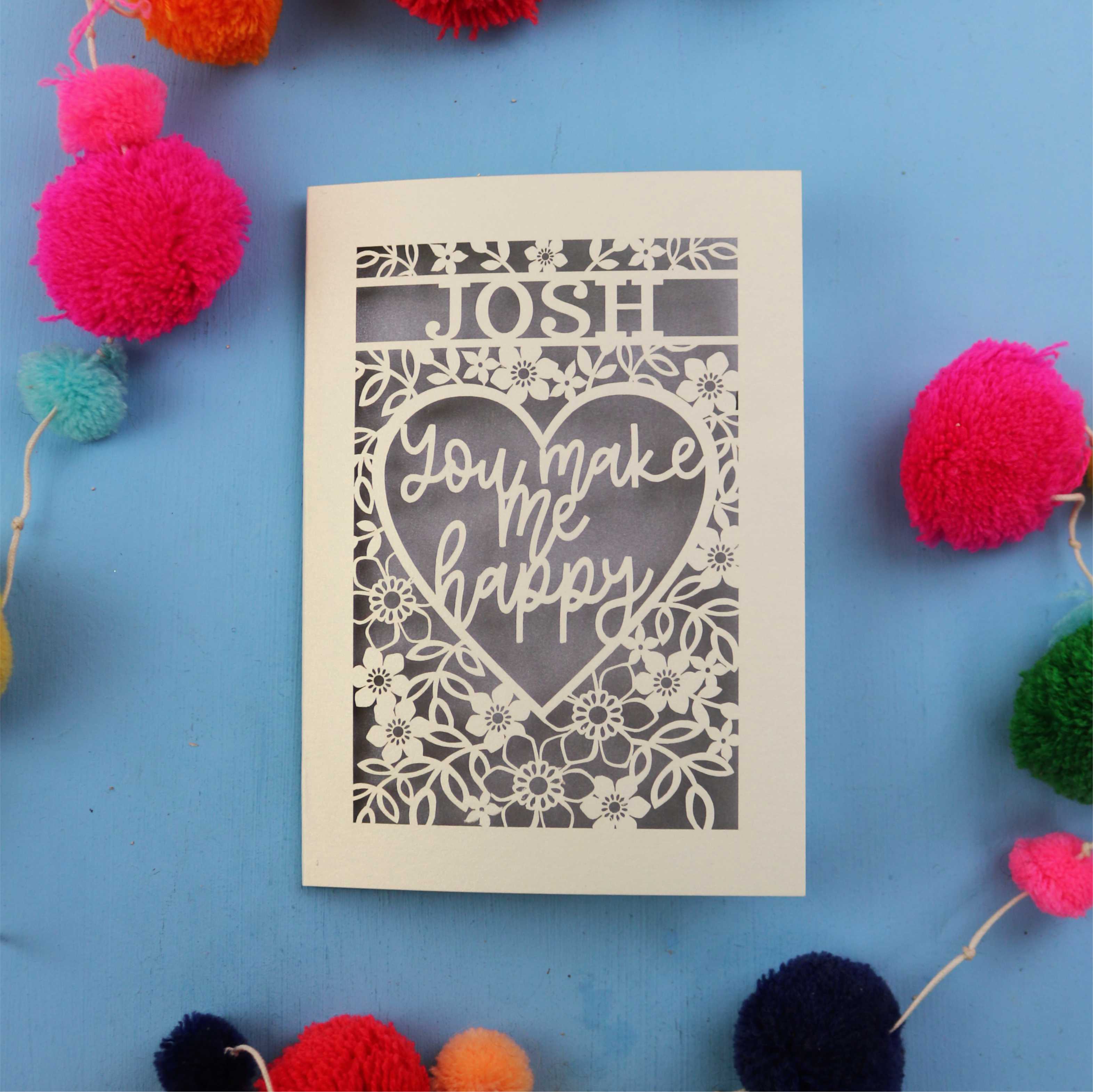 Laser cut Valentines Card with 'Josh you make me happy' text on a blue background with colourful pom-poms
