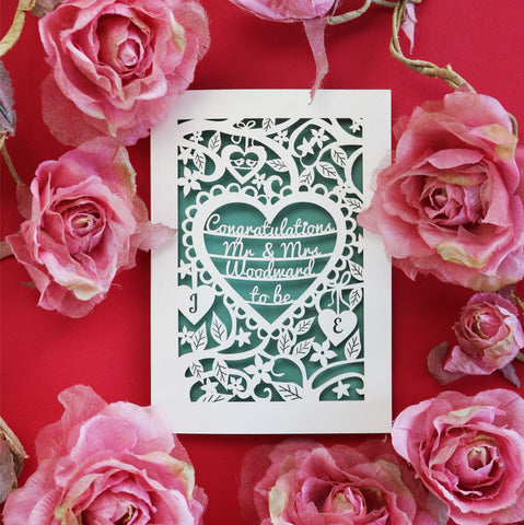A personalised engagement card, laser cut with floral details and a heart in the centre. Heart contains the words "Congratulations Mr and Mrs Woodward to be"
