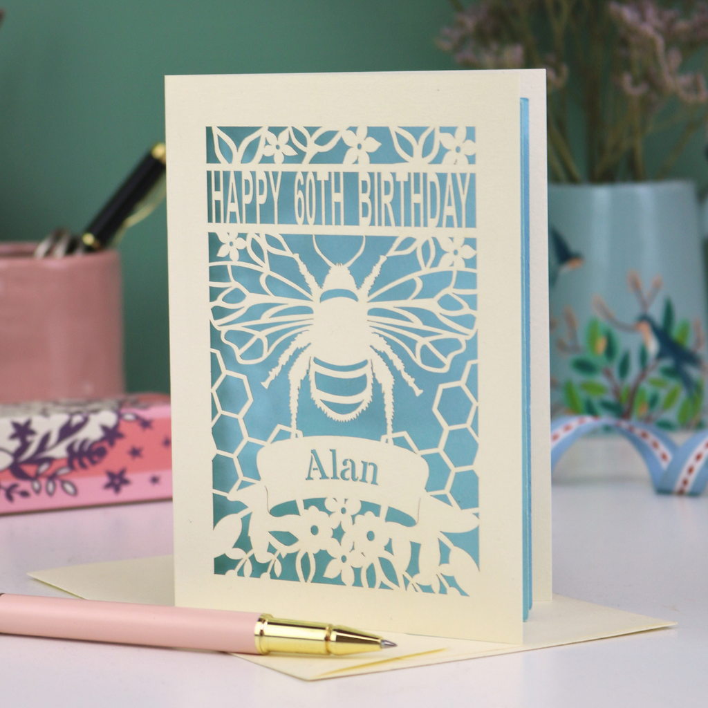 Birthday card with bee design and 'Happy 60th Birthday Alan' text on a desk with stationery items.
