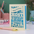A laser cut husband Birthday card for the most awesome husband ever with blue and cream cut out design on a desk with stationery items.