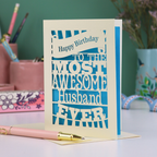 A laser cut husband Birthday card for the most awesome husband ever with blue and cream cut out design on a desk with stationery items.