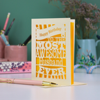 A laser cut husband Birthday card for the most awesome husband ever with yellow and cream cut out design on a desk with stationery items.
