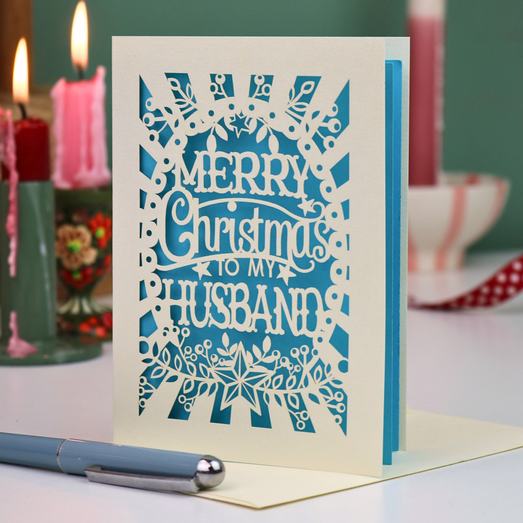 A laser-cut Christmas greeting card with the text 'Merry Christmas to my Husband' in cream cut out lettering over a blue background, standing on a desk with a pen in the foreground and blurred candles in the background.