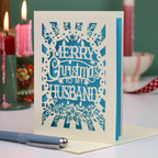 A laser-cut Christmas greeting card with the text 'Merry Christmas to my Husband' in cream cut out lettering over a blue background, standing on a desk with a pen in the foreground and blurred candles in the background.