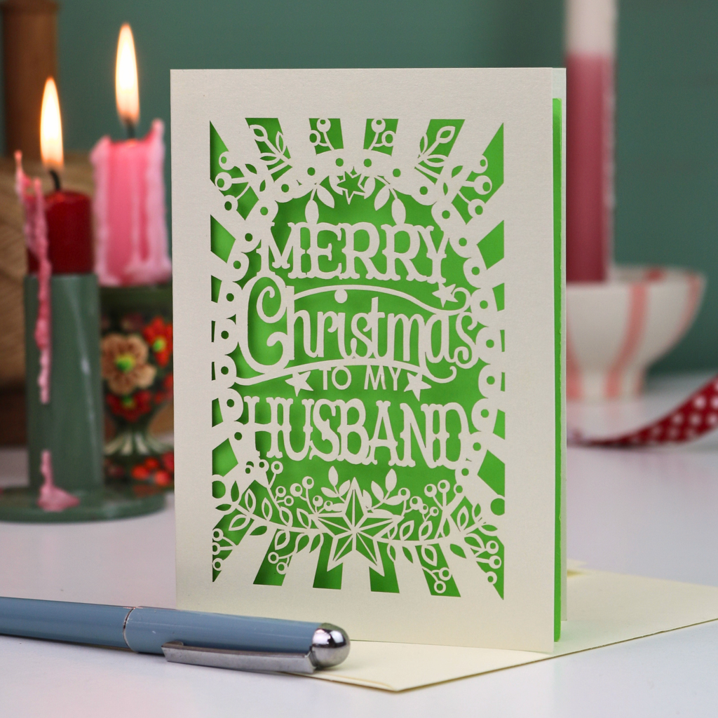 A laser-cut husband Christmas card with the text 'Merry Christmas to my Husband' in an ornate cut out design, displayed on a tabletop with candles and a pen in a softly blurred background.