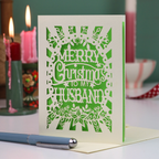 A laser-cut husband Christmas card with the text 'Merry Christmas to my Husband' in an ornate cut out design, displayed on a tabletop with candles and a pen in a softly blurred background.