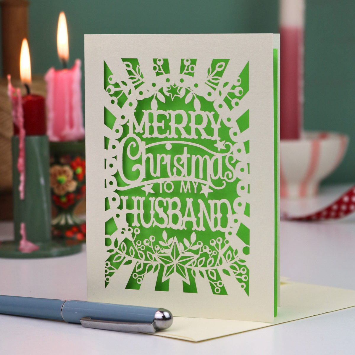 A laser-cut husband Christmas card with the text 'Merry Christmas to my Husband' in an ornate cut out design, displayed on a tabletop with candles and a pen in a softly blurred background.