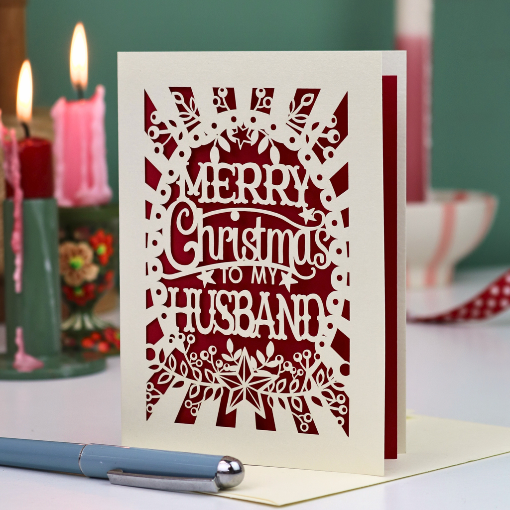 A laser-cut husband Christmas card with the text 'Merry Christmas to my Husband' in a paper cut out design, displayed on a tabletop with candles and a pen in a softly blurred background.
