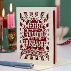 A laser-cut husband Christmas card with the text 'Merry Christmas to my Husband' in a paper cut out design, displayed on a tabletop with candles and a pen in a softly blurred background.