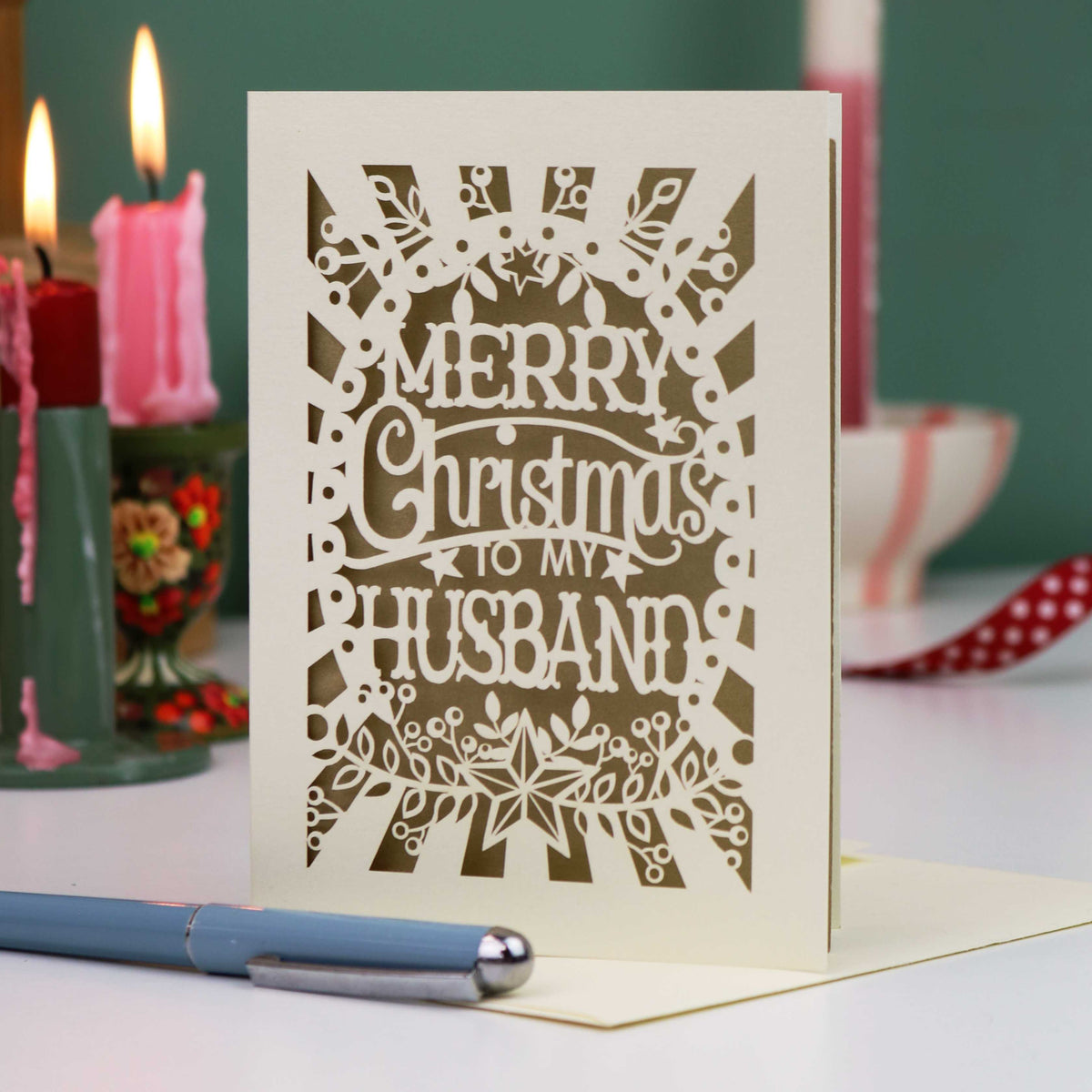 A laser-cut husband Christmas card with the text 'Merry Christmas to my Husband' in an ornate cut out design, displayed on a tabletop with candles and a pen in a softly blurred background.