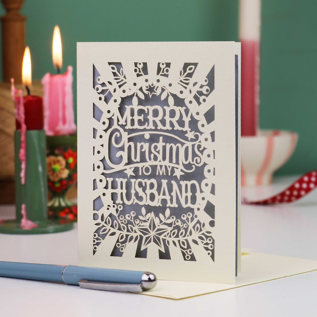 A laser-cut Christmas card with the text 'Merry Christmas To My Husband' in an intricate snowflake and floral design, displayed on a table with candles in the background.